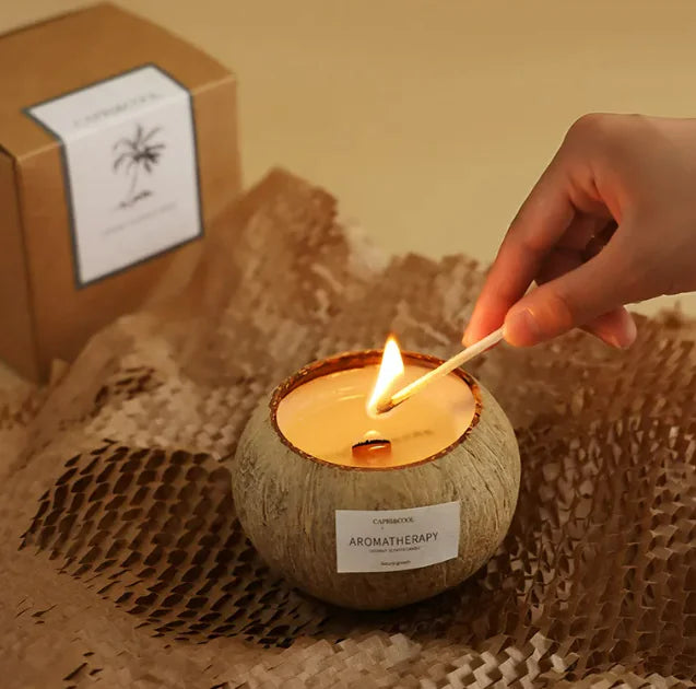 Person lighting an aromatherapy coconut candle in a natural coconut shell, with a gift box in the background.